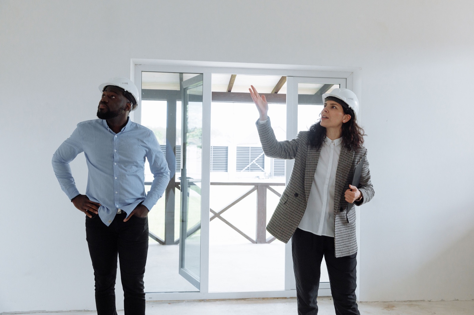 A woman in a hard hat holding a folder points out details to a man in a hard hat inside an unfinished room with glass doors