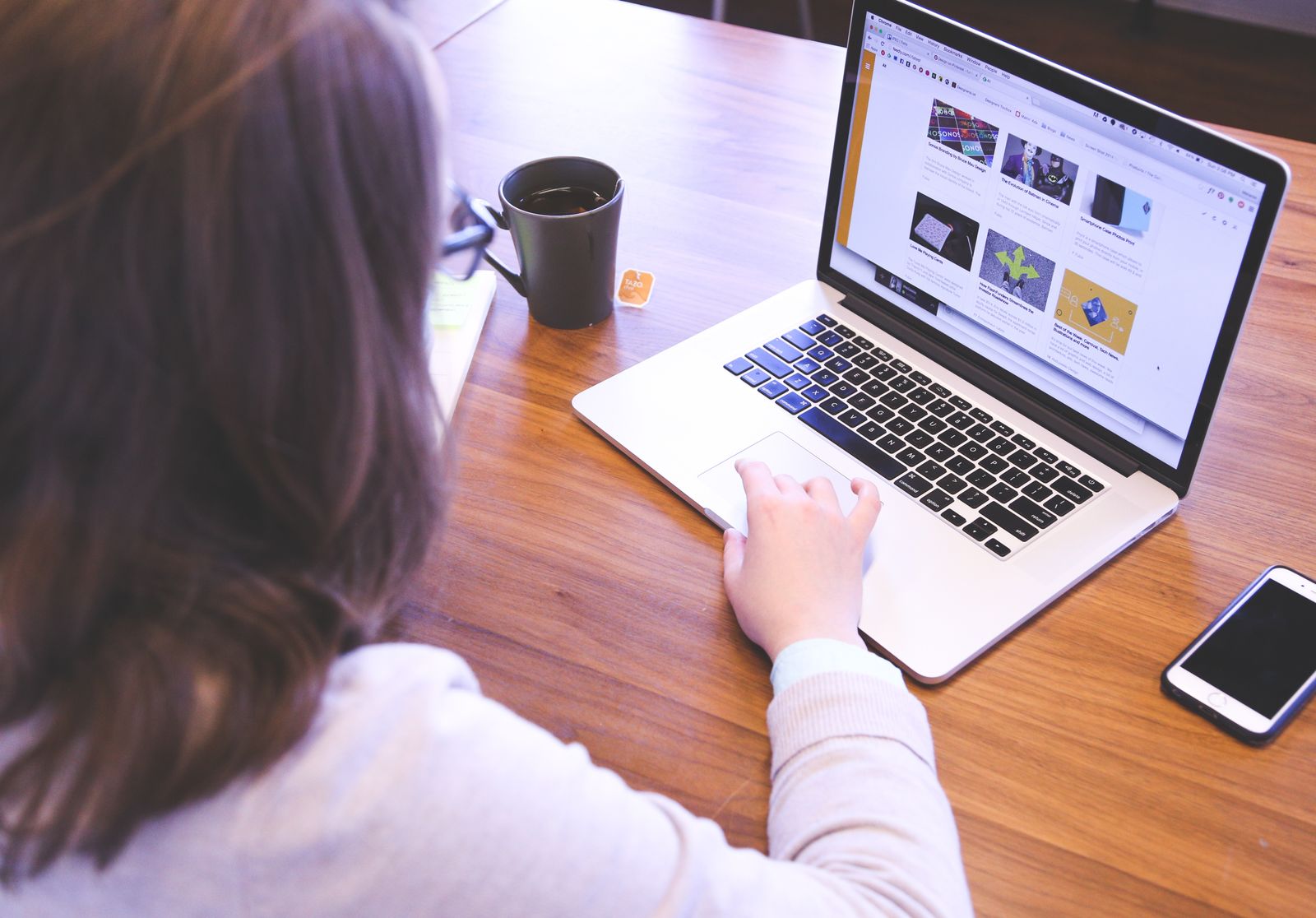 A person browsing a website on a laptop at a wooden desk, with a coffee mug and a phone nearby