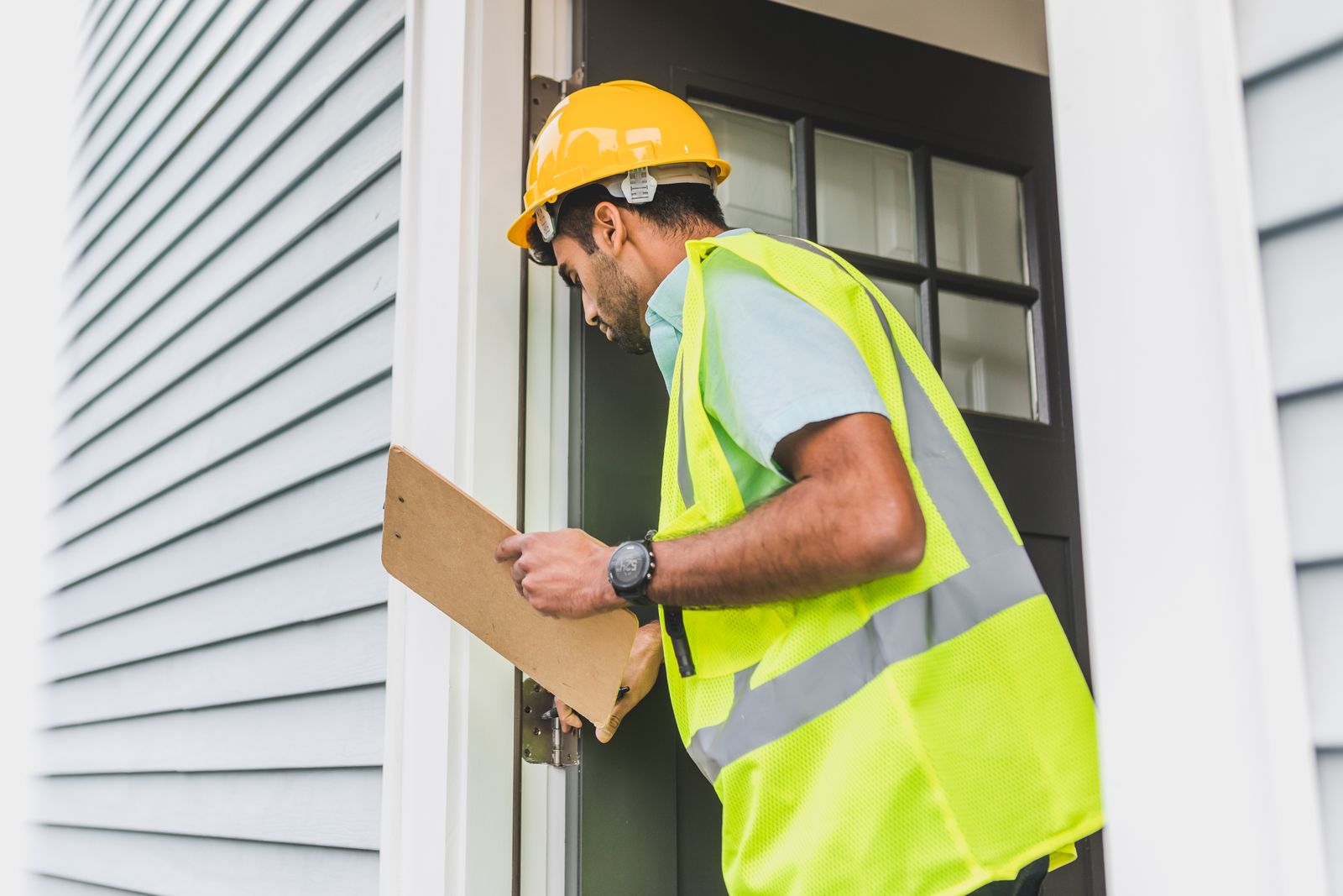 A contractor in a hard hat and safety vest inspecting a front door, clipboard in hand