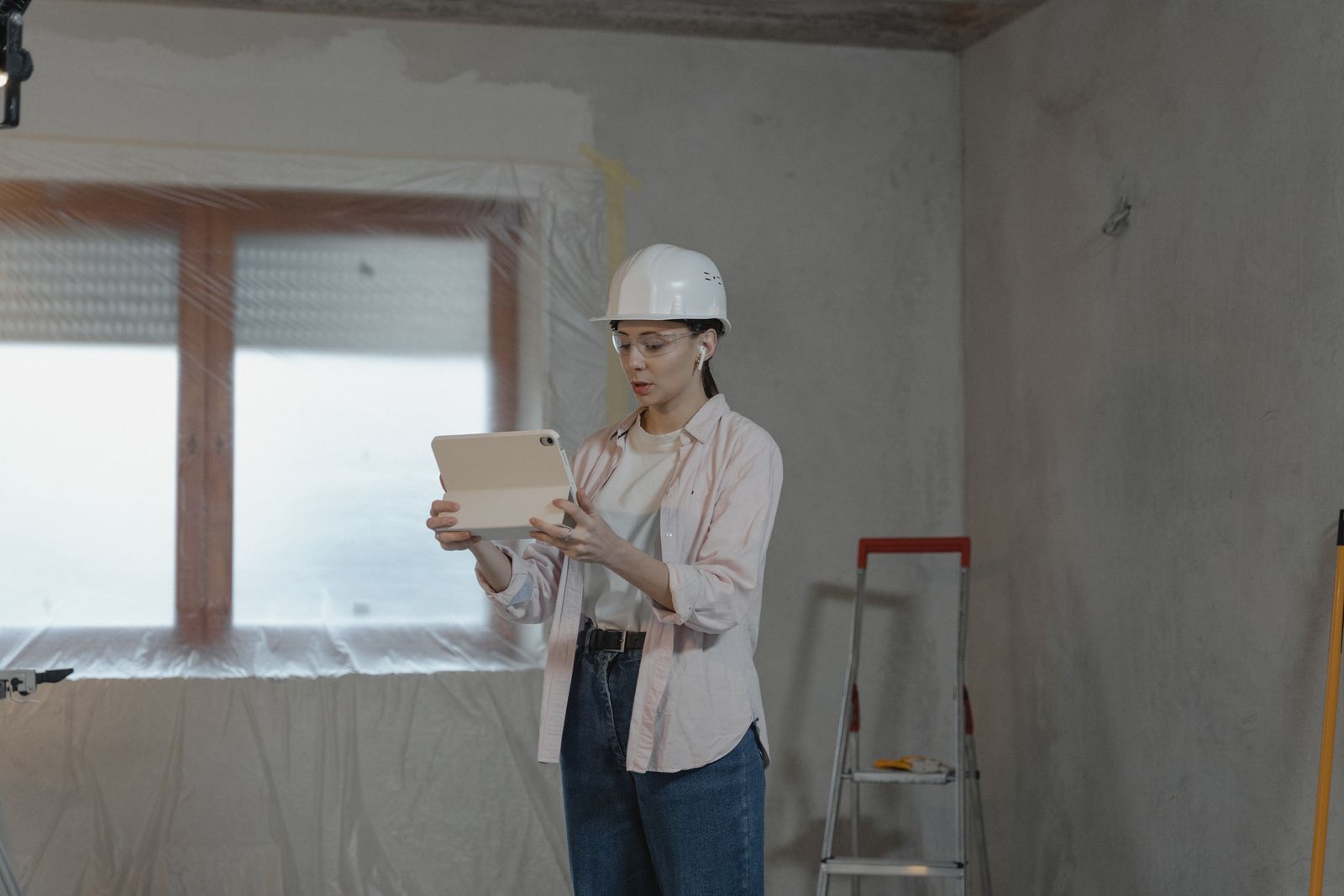 A woman in a hard hat and safety glasses reviewing a tablet in a room under renovation