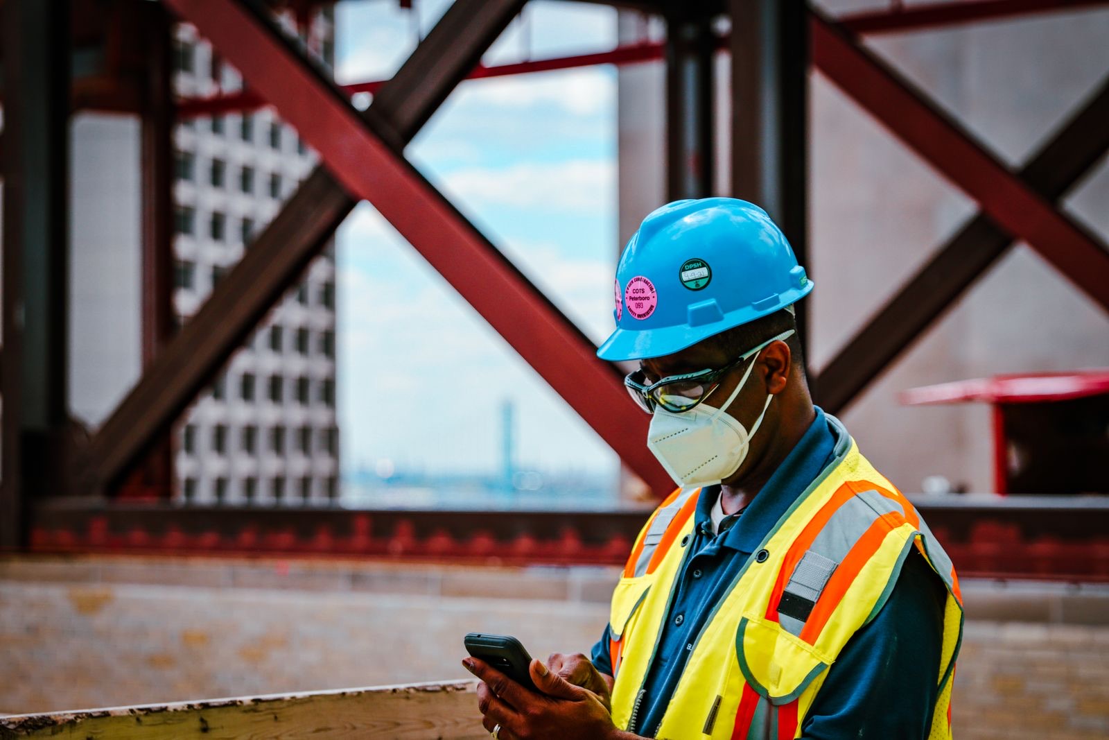 A worker in a blue hard hat, safety glasses, mask and high-visibility vest looking at a phone on a building site with steel beams behind him