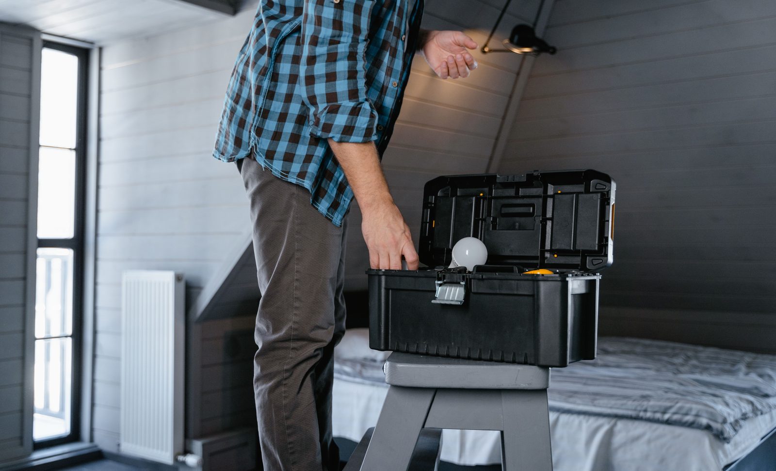 A person in a plaid shirt opening a toolbox on a stepladder in an attic bedroom, holding a light bulb