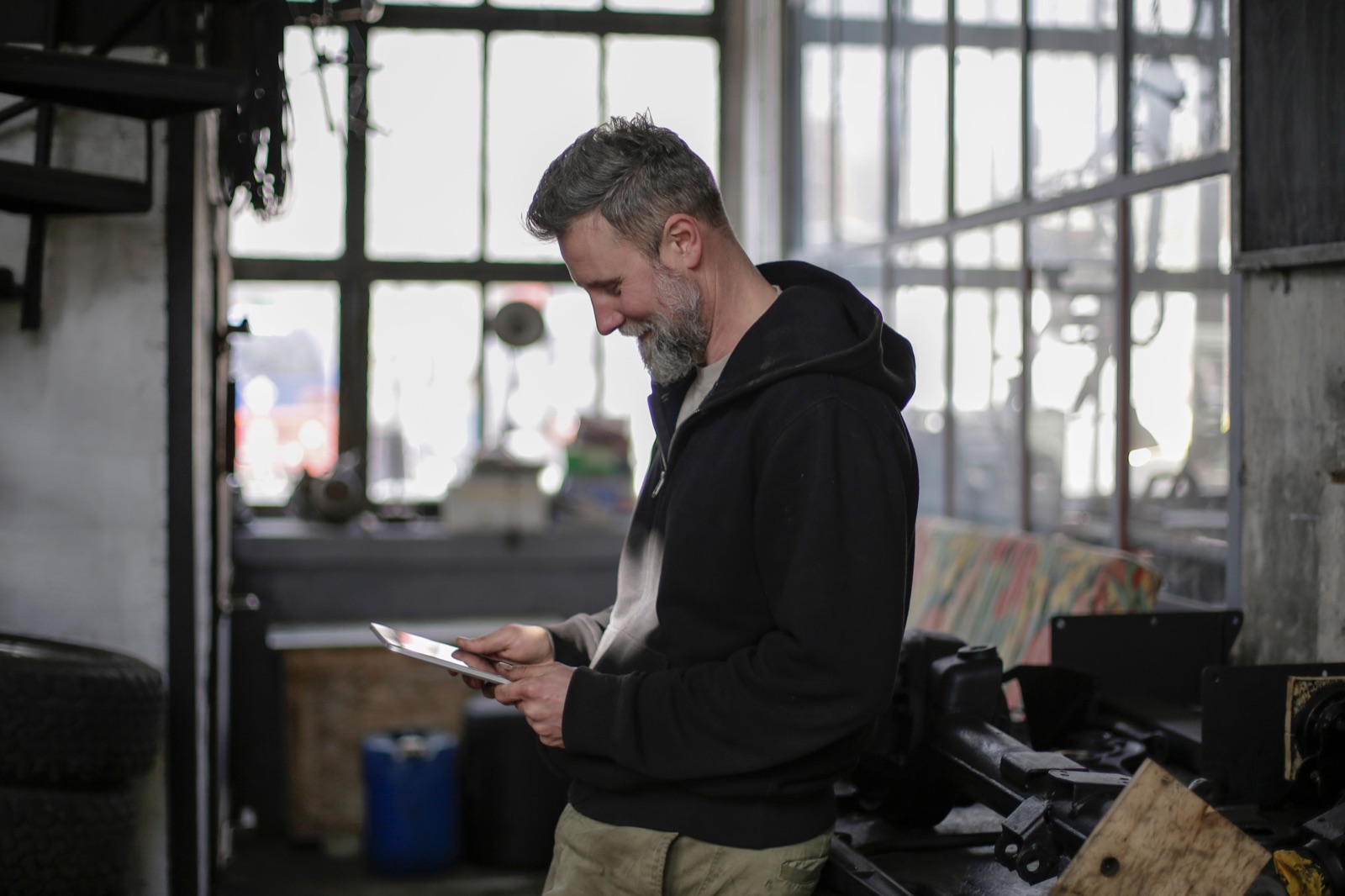 A bearded worker in a hoodie smiling at a tablet in a workshop