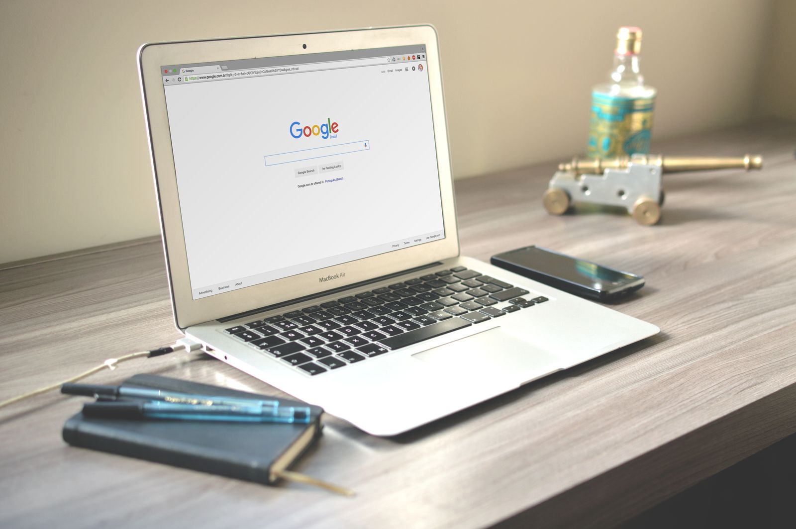 A laptop showing the Google search page on a wooden desk next to a phone and a notebook