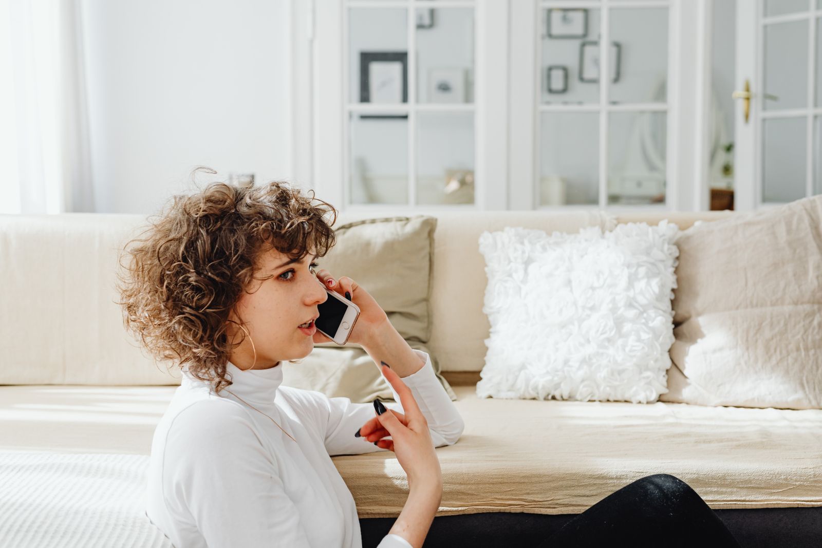 A woman sitting by a sofa and talking on her phone at home