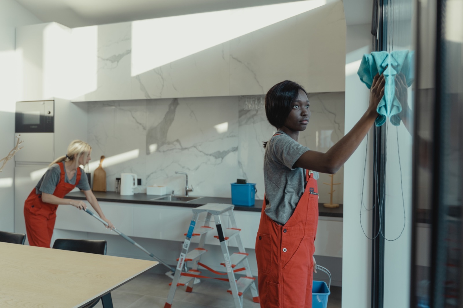 Two cleaners in orange overalls working in a modern kitchen, one wiping a glass door with a blue cloth while the other mops the floor