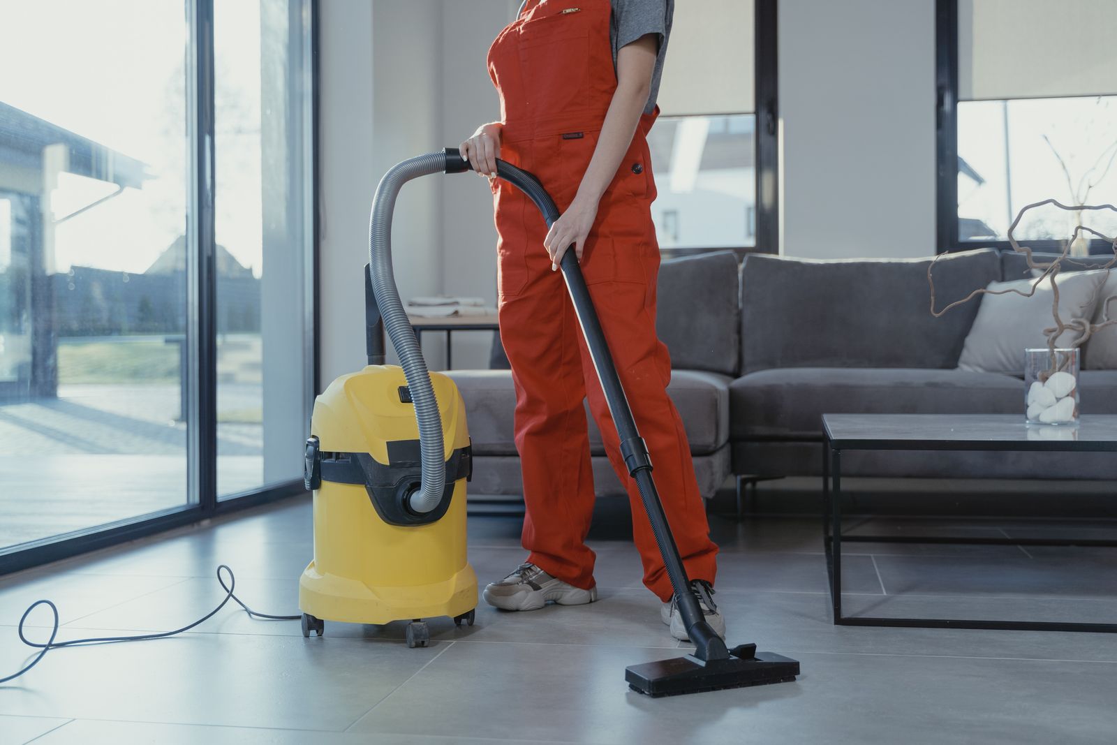 A cleaner in red overalls vacuuming the tiled floor of a bright living room with a yellow canister vacuum