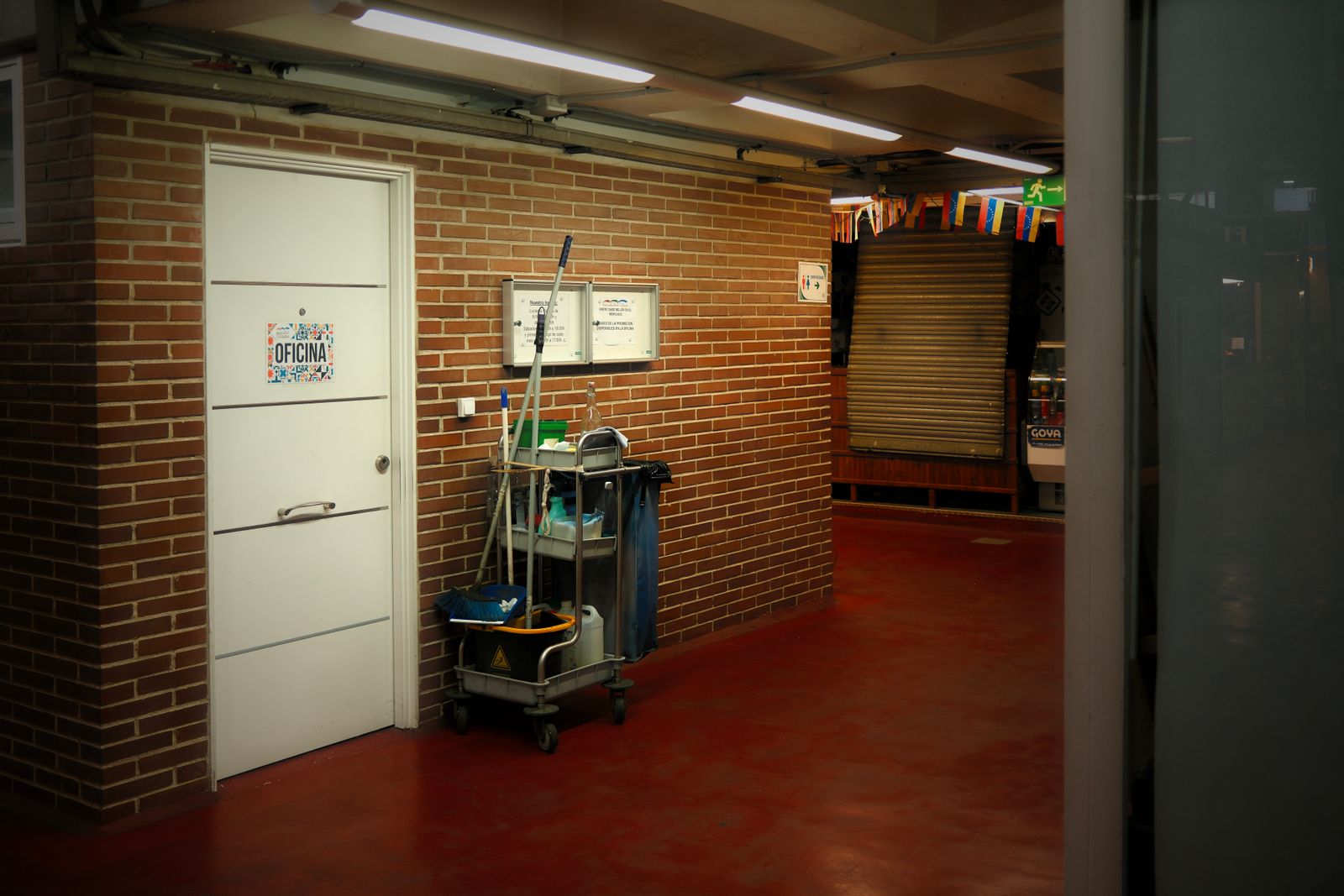 A janitorial cart with a mop, bucket and supplies parked in a brick hallway next to an office door marked 'Oficina'