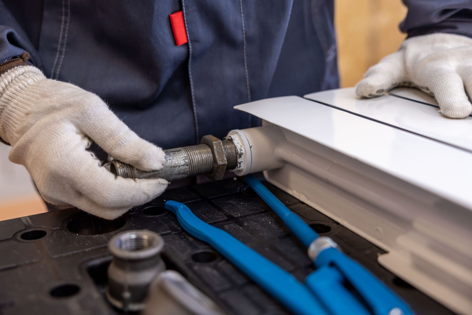 Gloved hands threading a steel pipe fitting onto a radiator, with blue pipe wrenches on the workbench