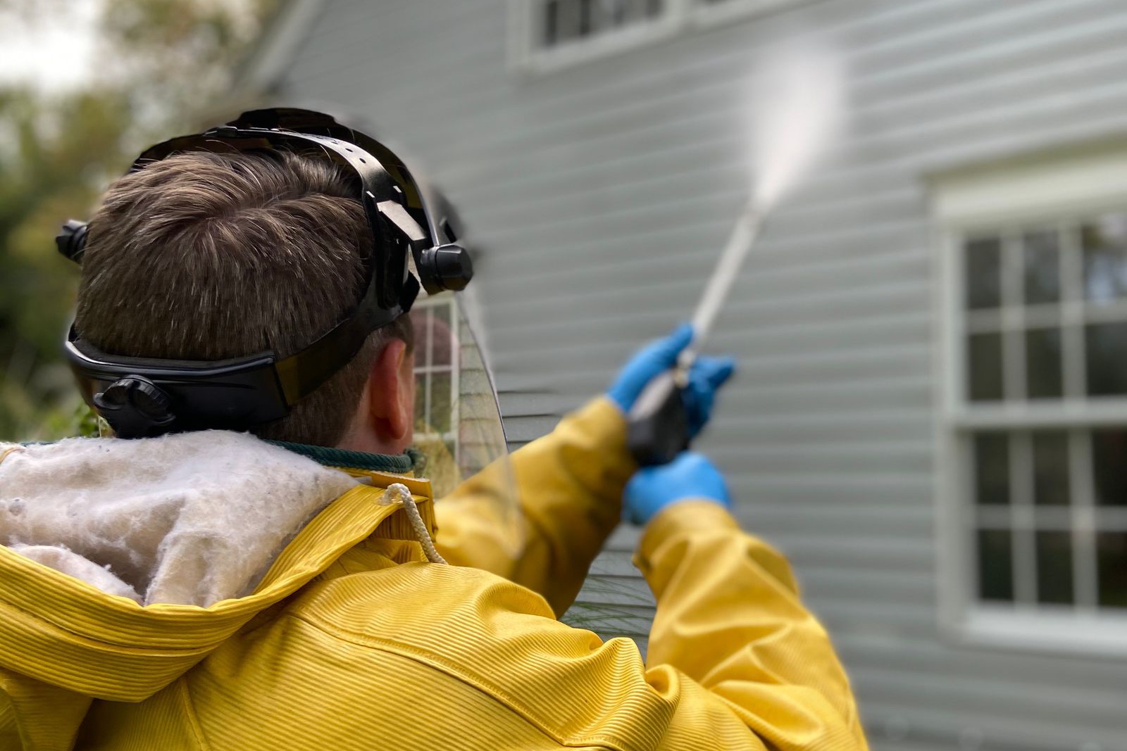 A worker in a yellow rain jacket, face shield and gloves spraying the vinyl siding of a house with a pressure washer wand