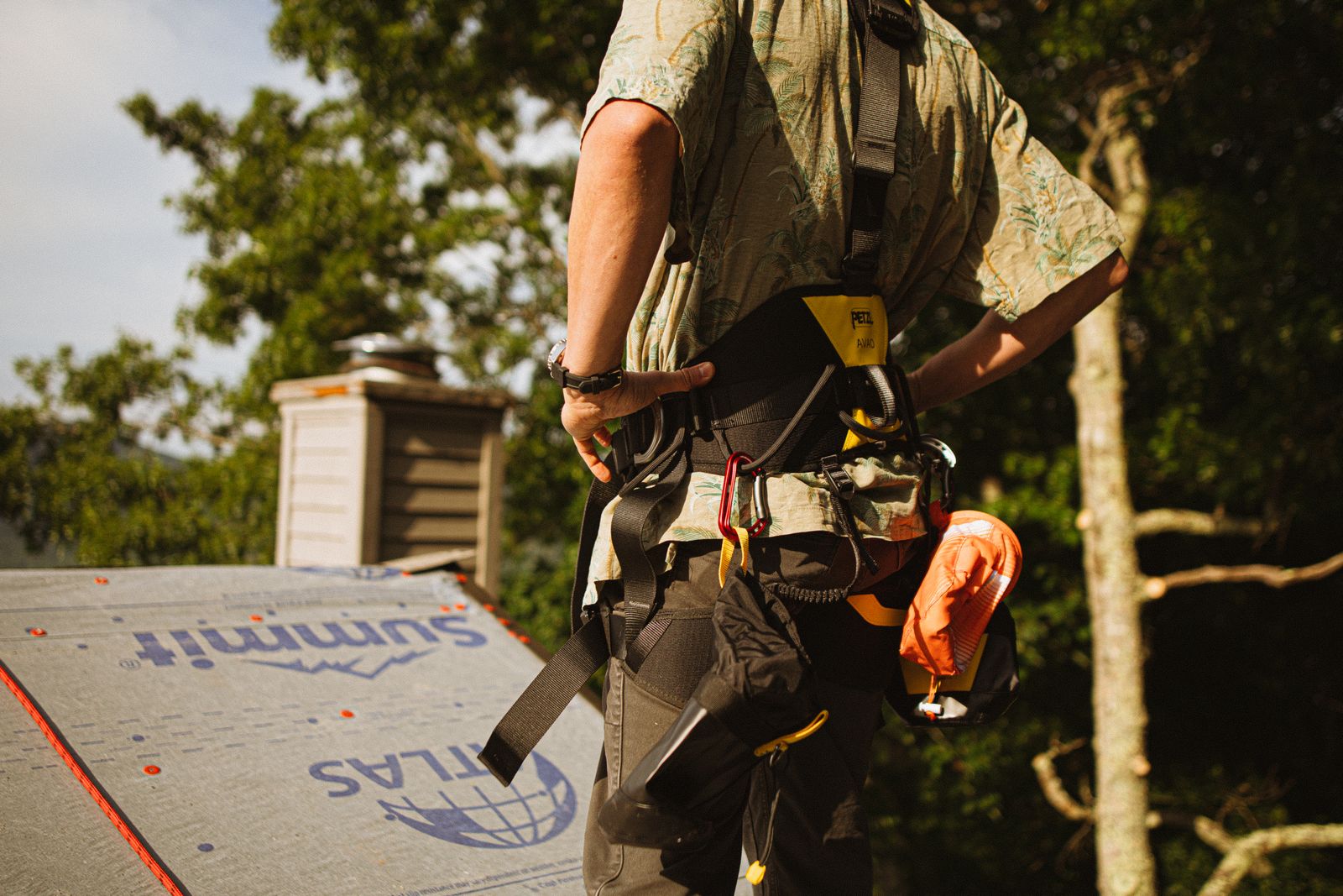 A roofer wearing a full-body fall protection harness with carabiners and a tool pouch, beside a roof covered in synthetic underlayment