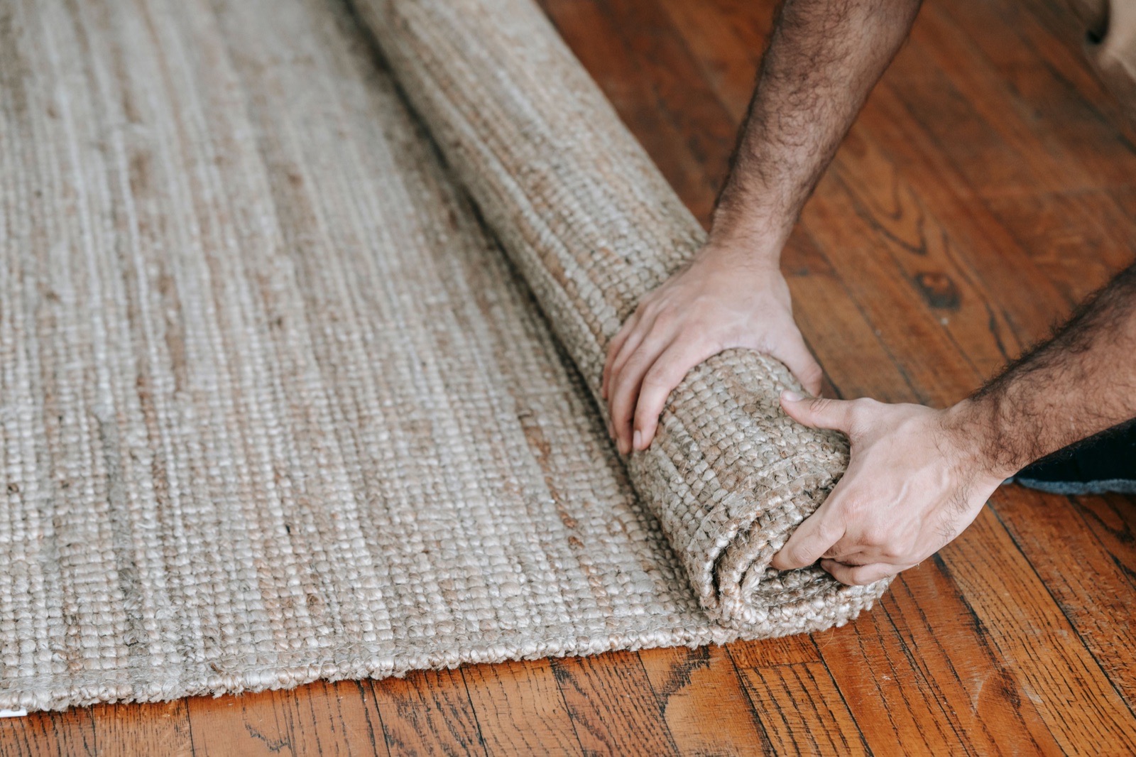 Two hands rolling up a woven area rug on a wooden floor