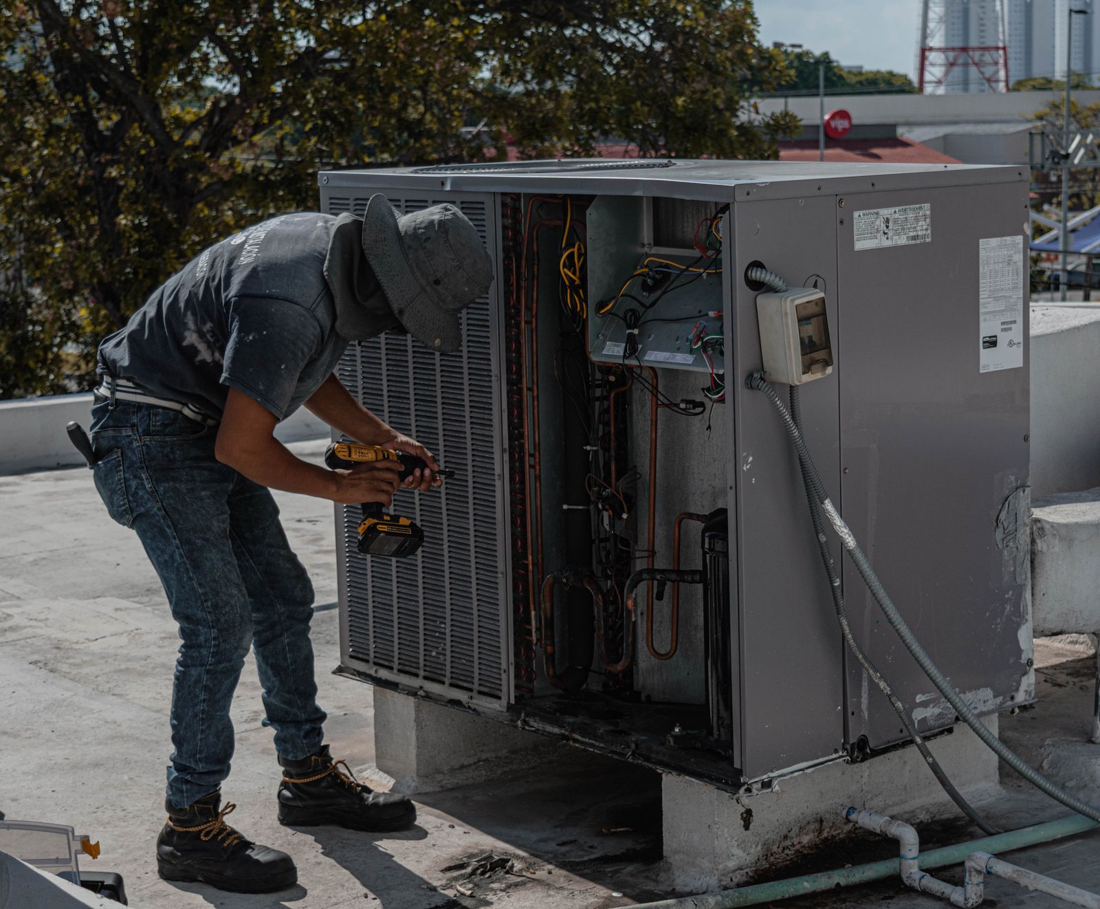 An HVAC technician using a cordless drill on an open rooftop condensing unit, its wiring and copper piping exposed