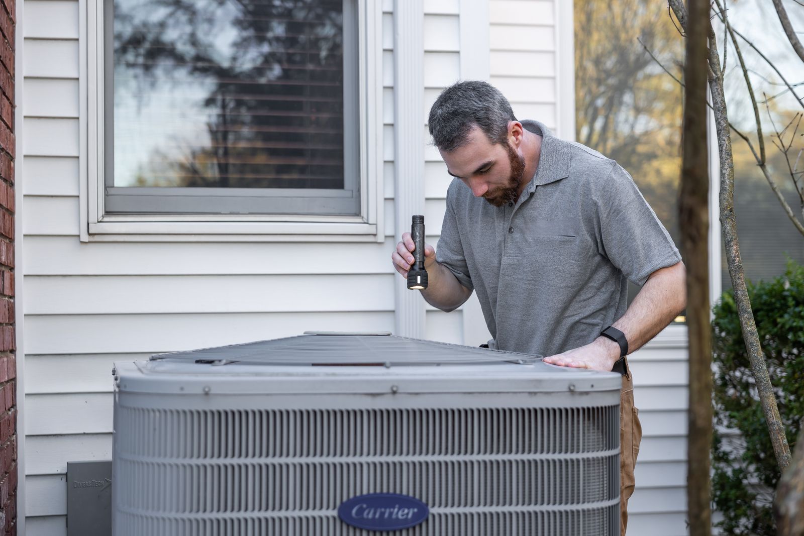 An HVAC technician inspecting an outdoor air conditioning unit beside a house with a flashlight