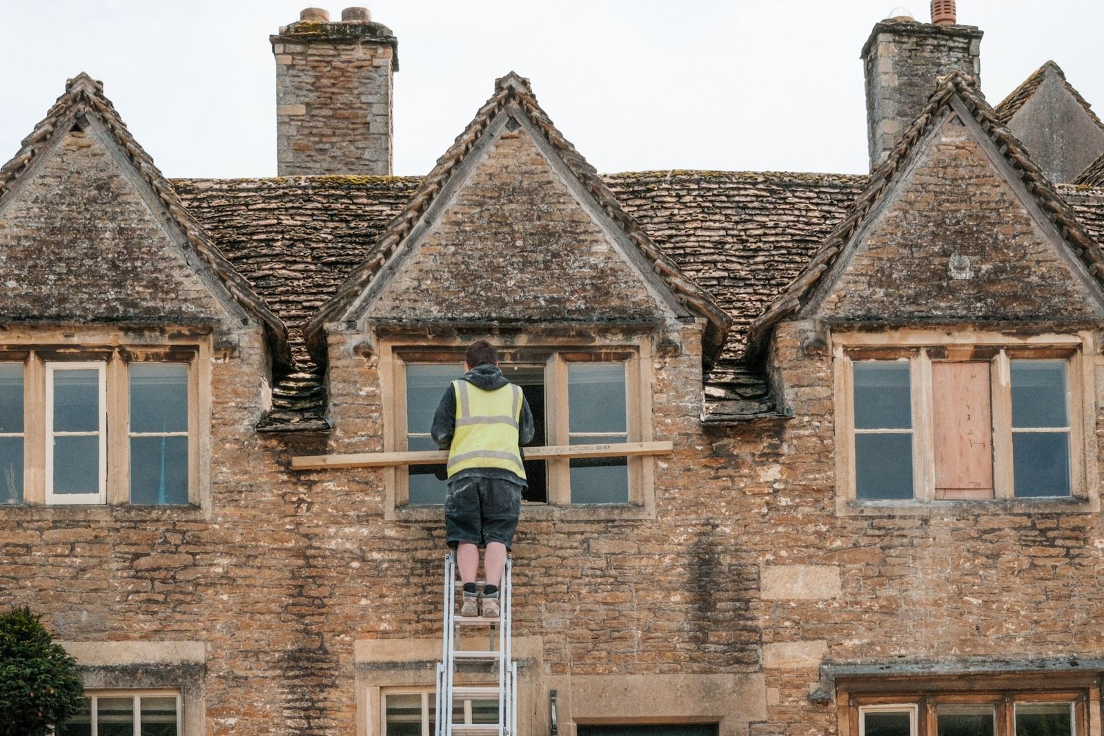 A worker in a high-visibility vest on a ladder repairing a window on a stone house