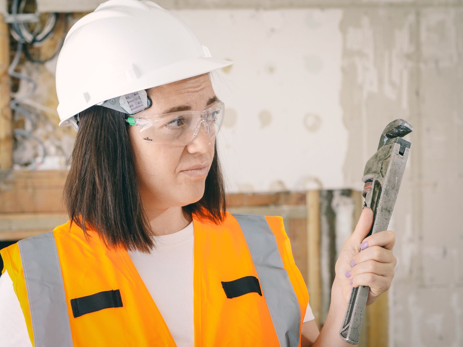 A tradeswoman in a hard hat and safety glasses holding a large pipe wrench