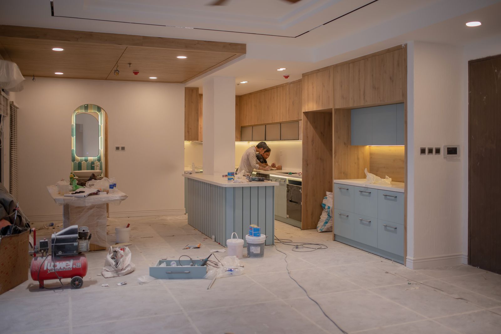 Two workers at the island of an open-plan kitchen nearing the end of a remodel, with tools and supplies on the floor