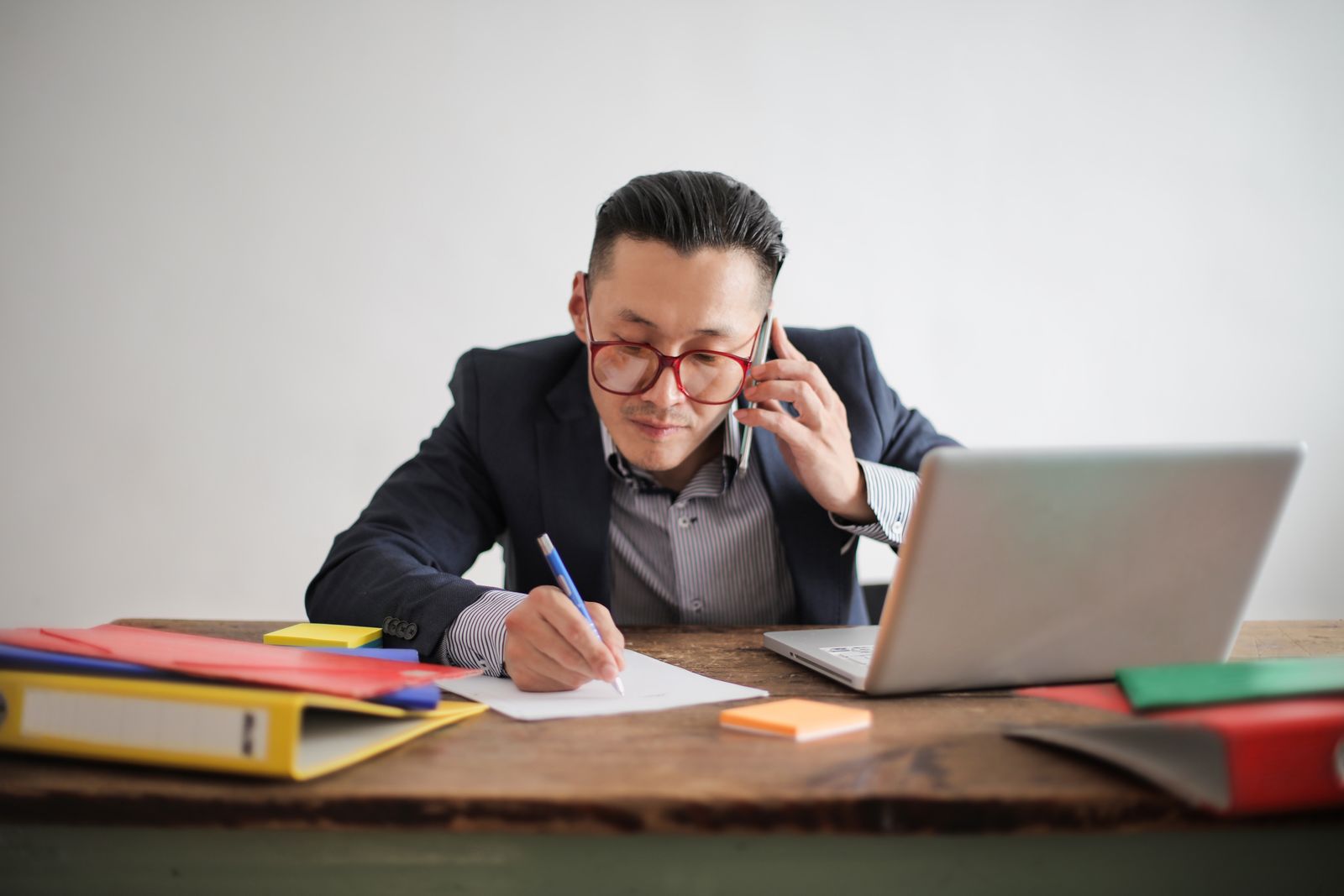 A man on a phone call taking notes at a desk with a laptop