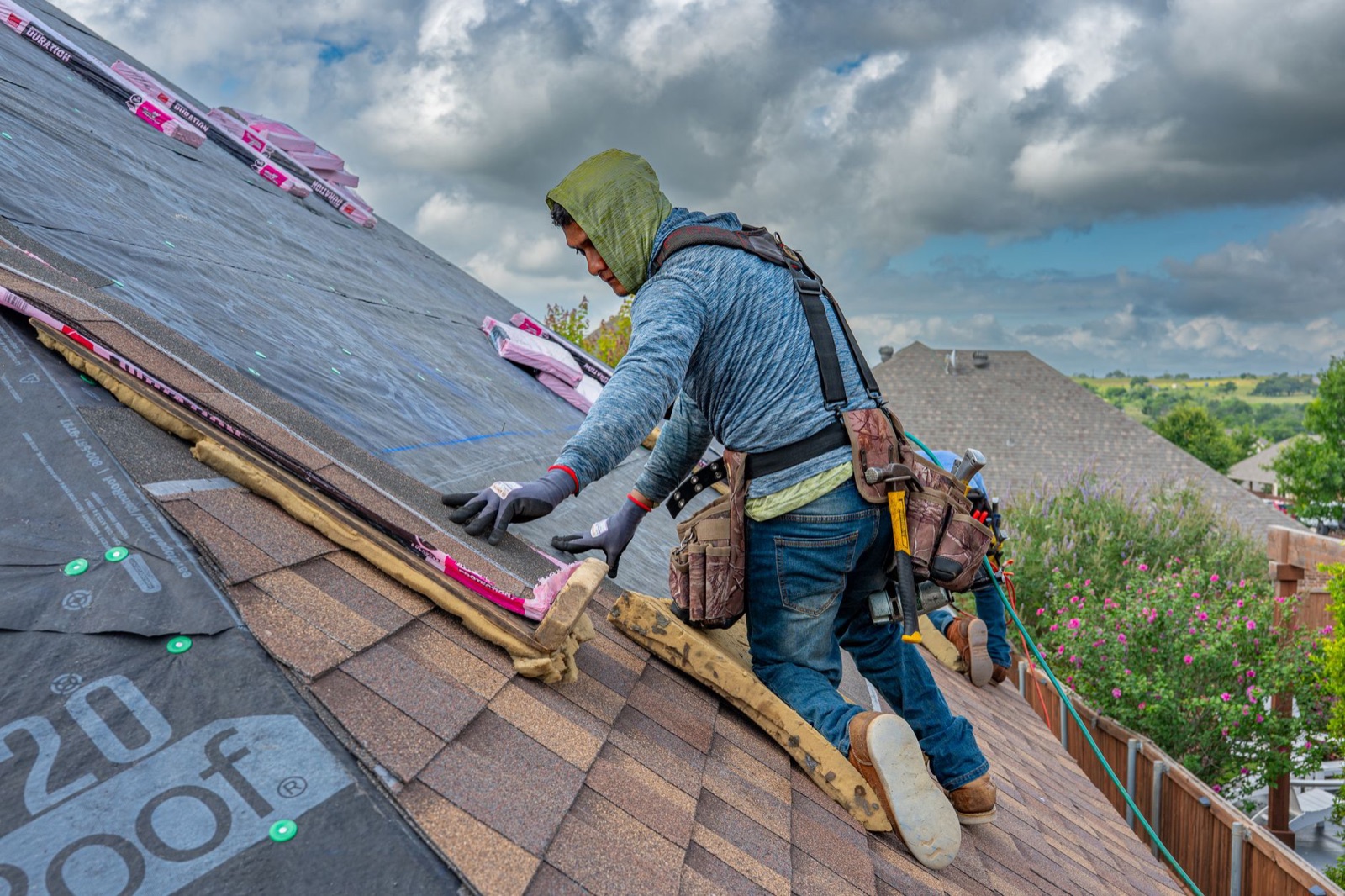A roofer in a tool belt and harness laying asphalt shingles on a steep roof under a cloudy sky, with another worker behind him
