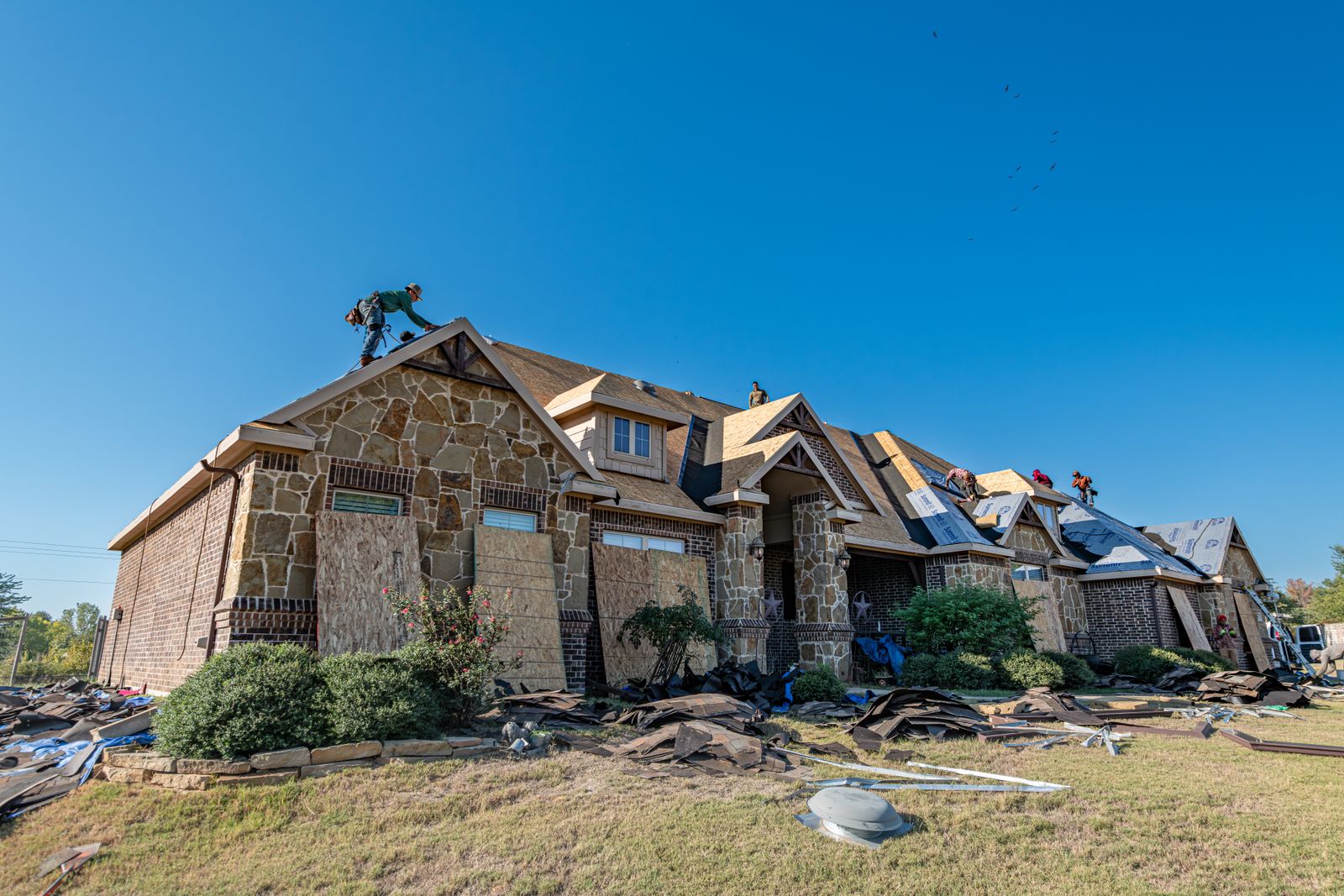 Roofers working on the roof of a brick and stone house, with old shingles on the lawn